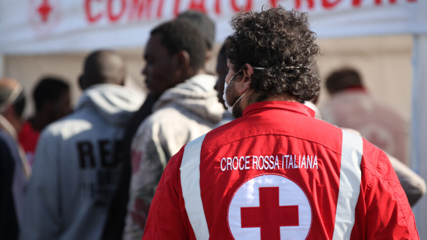 A member of the Italian red cross stands on a pier next to a group of migrants