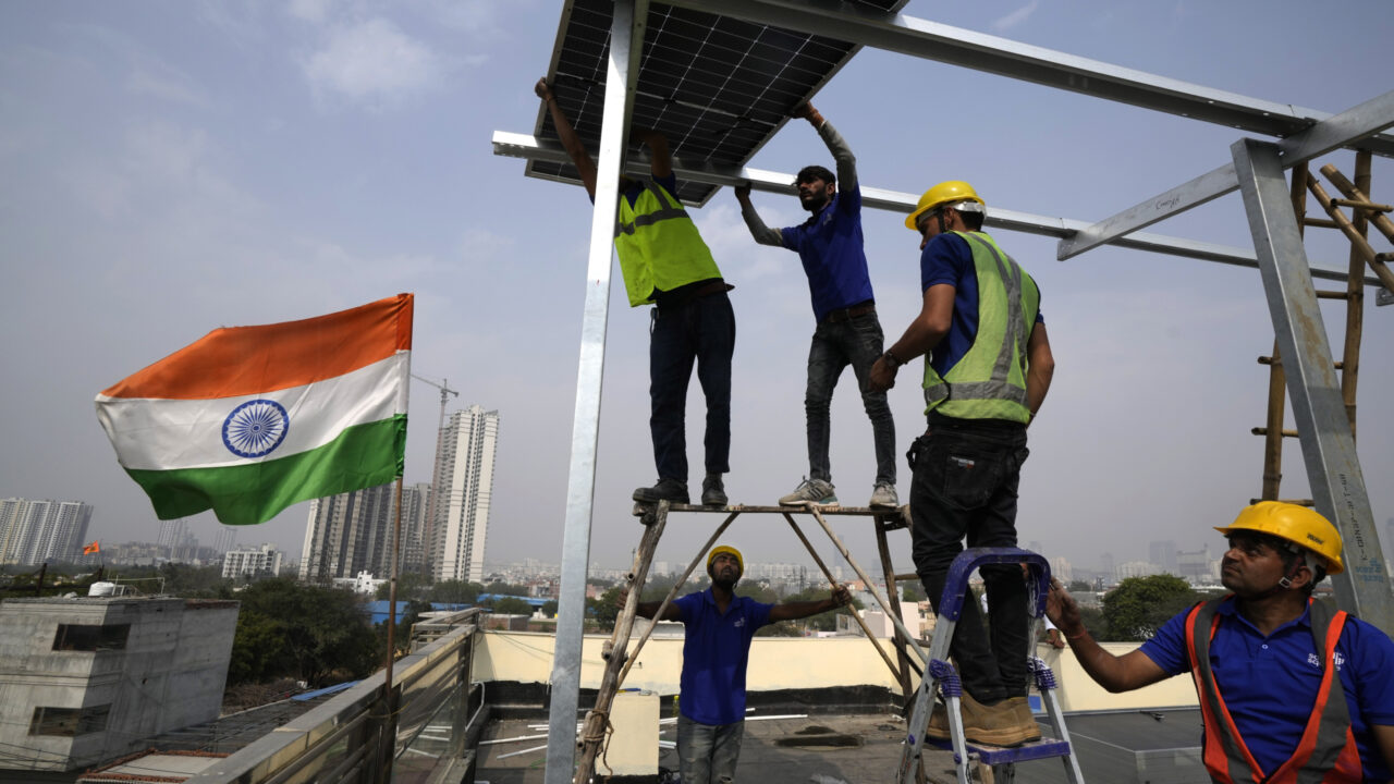 Four workers in hi-vis vests place a solar panel on the rooftop of a residence in Gurugram on the outskirts of New Delhi, India, as an Indian flag flutters to the left