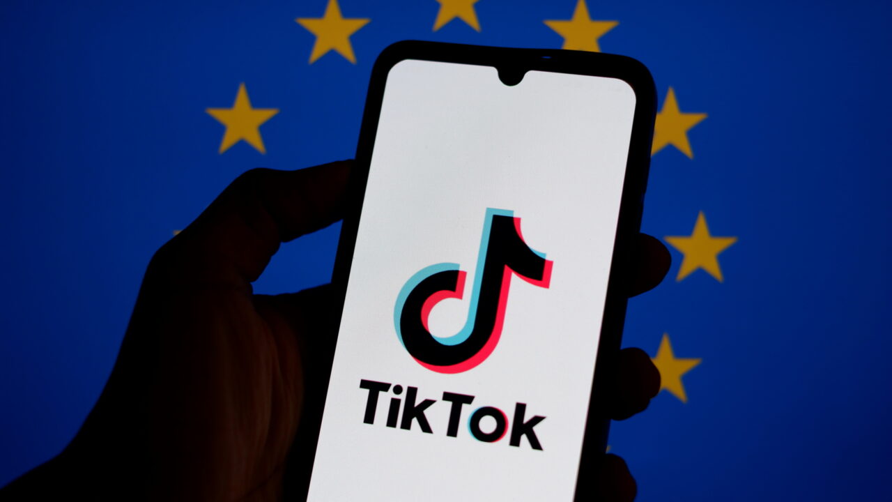 Hand holding a phone with the TikTok logo on the screen. EU flag is in the background