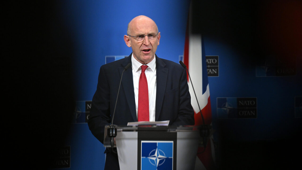 British Defense Secretary John Healey standing behind a podium with the words NATO, and the accompanying NATO symbol, at a press conference following the Ukraine Defense Contact Group meeting held at NATO Headquarters in Brussels. A British Flag hangs to his right