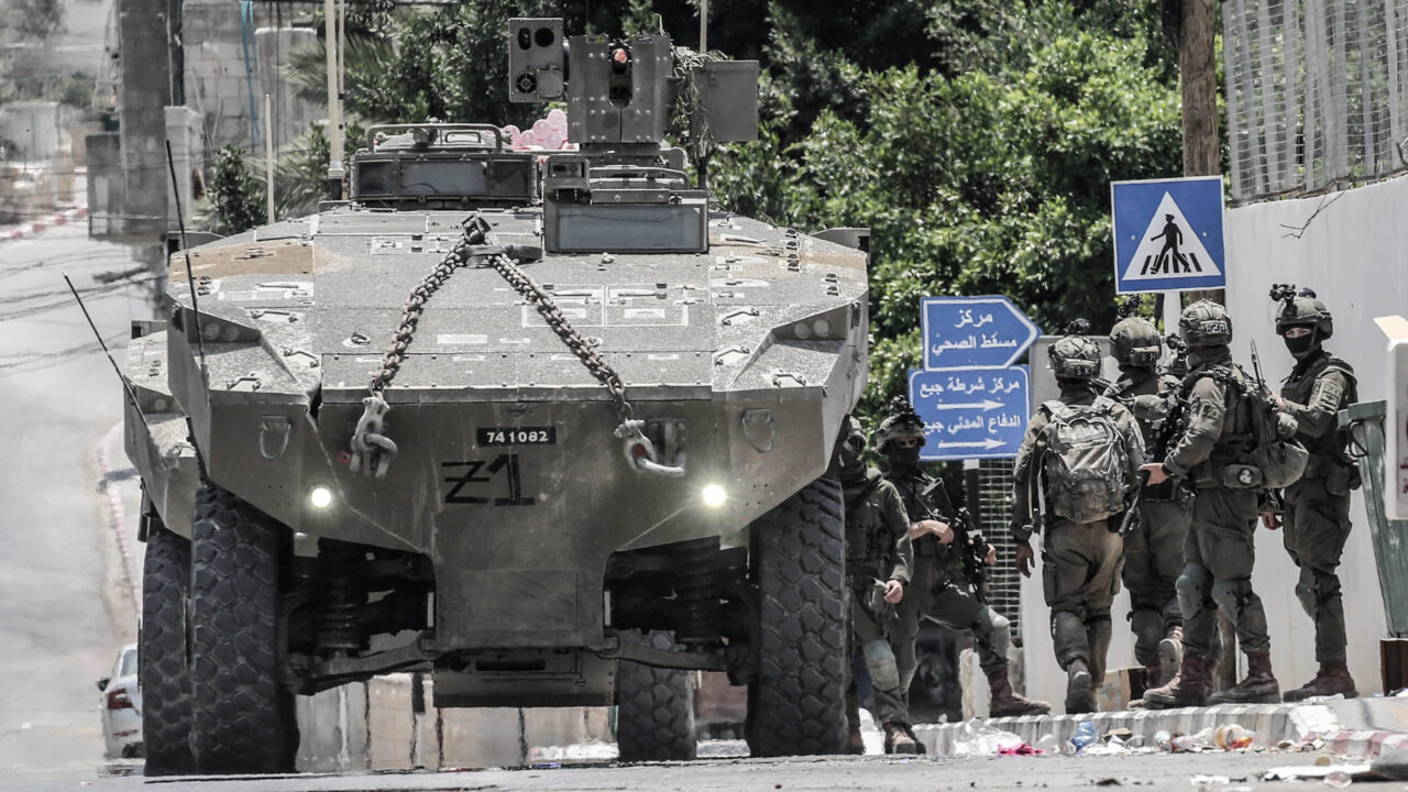 Israeli Occupation Forces (right) stand next to an armoured vehicle (left) on a street with arabic road signs in the background
