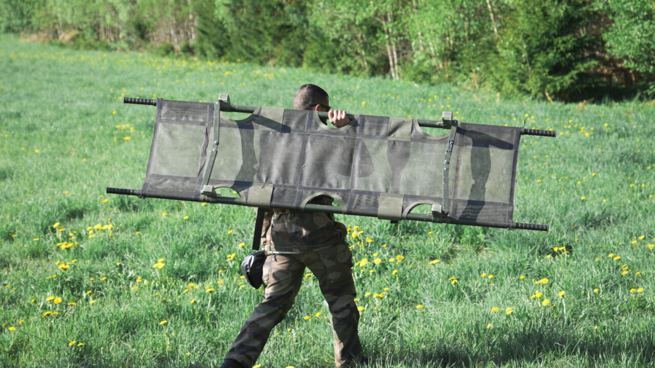 Soldier with back turned to the camera walks through a green field with a khaki green stretcher on his back