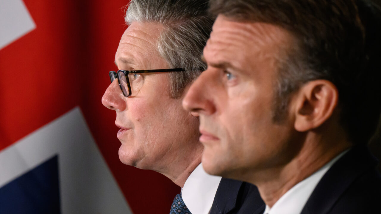 A closeup of Keir Starmer and Emmanuel Macron’s profiles as they look off to the left. A british flag hangs in the background