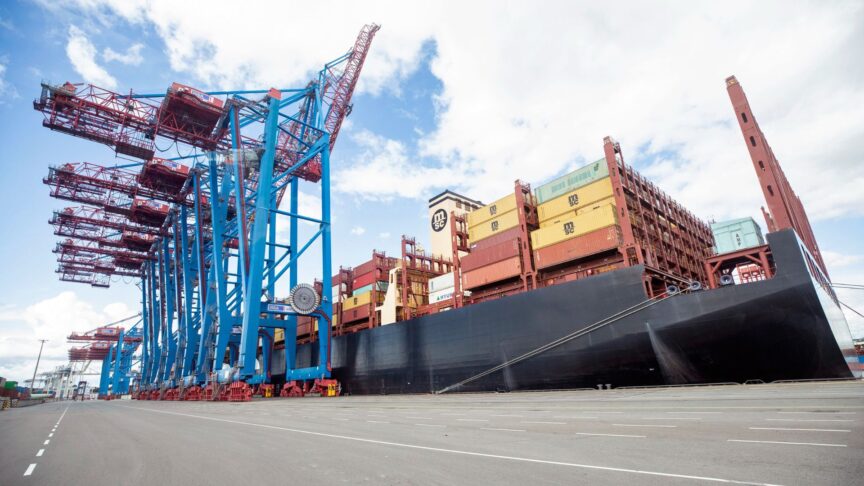 A large container ship loaded with containers is anchored at a port. To the left stands a row of cranes used to load and unload cargo
