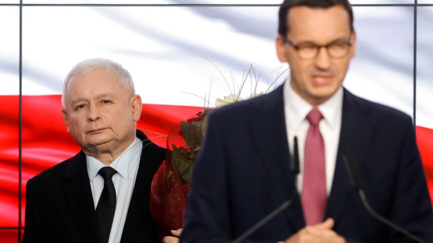 Ein Foto zeigt zwei Männer in Anzügen, einer am Rednerpult, im Hintergrund ein Mann mit Blumen vor einer rot-weißen Flagge.
