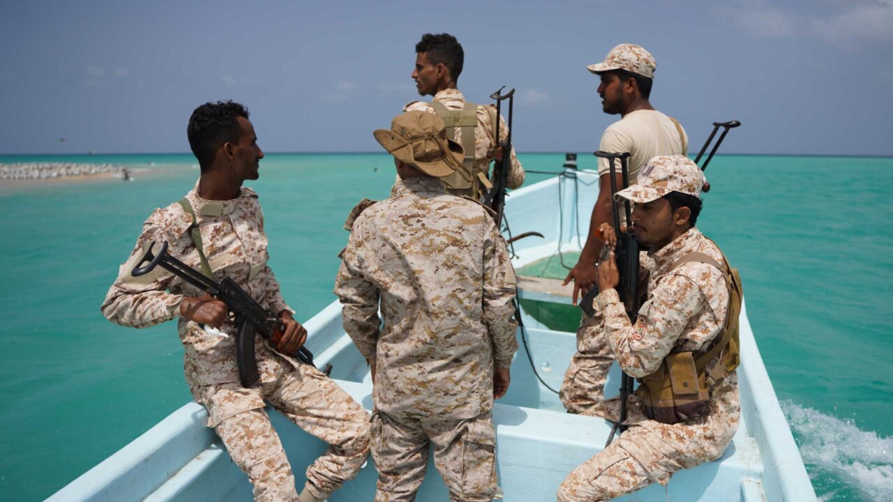 Ein Foto zeigt fünf Soldaten in hellen Wüstentarnuniformen mit Gewehren auf einem blauen Boot auf türkisfarbenem Meer