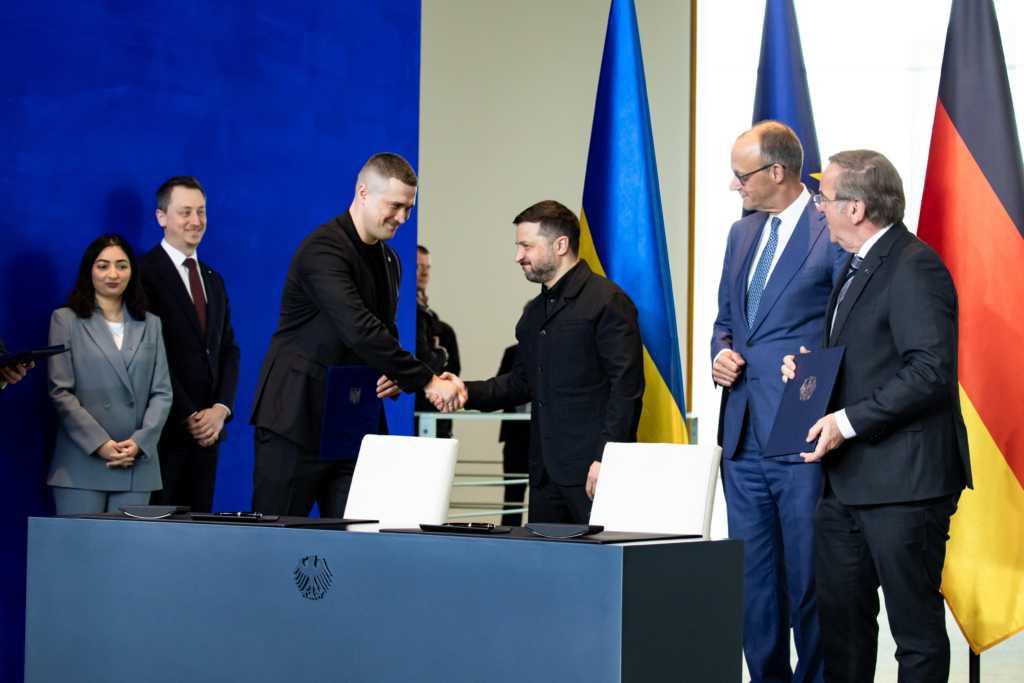 Ein Foto zeigt zwei Männer beim Händedruck vor einem Tisch in einem Innenraum mit ukrainischer und deutscher Flagge.
