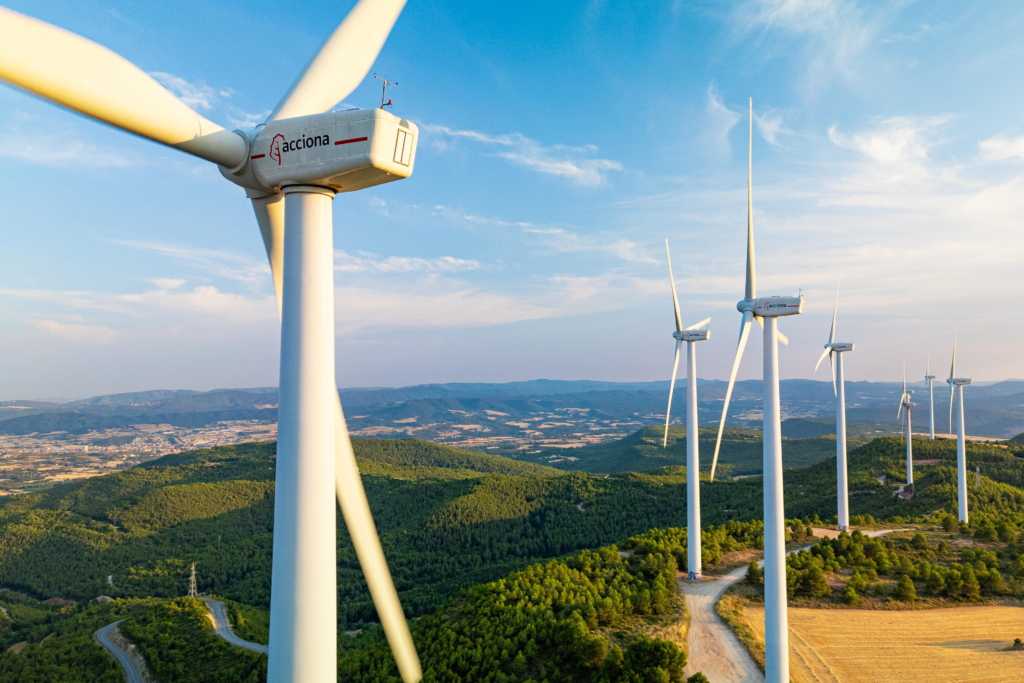 Ein Foto zeigt Windturbinen, die auf bewaldeten Hügeln in einer Windfarm stehen, aufgenommen aus der Luft bei klarem Himmel.