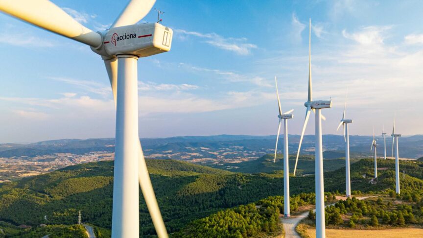 Ein Foto zeigt Windturbinen, die auf bewaldeten Hügeln in einer Windfarm stehen, aufgenommen aus der Luft bei klarem Himmel.