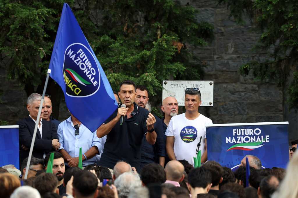 Ein Foto zeigt einen Mann am Mikrofon, der auf einem Podium vor einer Menschenmenge spricht, mit blauer Fahne und Bannern im Hintergrund.