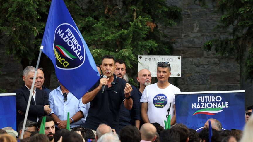 Ein Foto zeigt einen Mann am Mikrofon, der auf einem Podium vor einer Menschenmenge spricht, mit blauer Fahne und Bannern im Hintergrund.