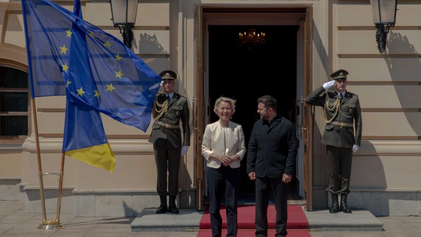 Ein Foto zeigt zwei Personen nebeneinander auf einem roten Teppich vor einem Gebäude, flankiert von Soldaten und Flaggen.
