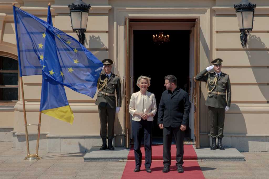 Ein Foto zeigt zwei Personen nebeneinander auf einem roten Teppich vor einem Gebäude, flankiert von Soldaten und Flaggen.