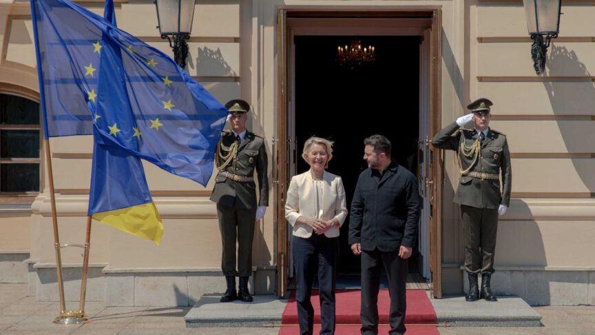 Ein Foto zeigt zwei Personen nebeneinander auf einem roten Teppich vor einem Gebäude, flankiert von Soldaten und Flaggen.