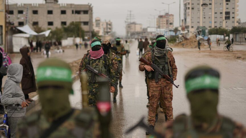 Ein Foto zeigt maskierte Kämpfer in militärischer Kleidung und grünen Stirnbändern, bewaffnet mit Gewehren, die in einer nassen Straße vor Wohngebäuden stehen.