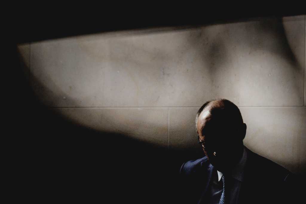 Ein Foto zeigt eine Person in dunklem Anzug mit gesenktem Kopf vor einer hellen Wand und starken Schatten.