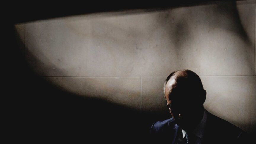 Ein Foto zeigt eine Person in dunklem Anzug mit gesenktem Kopf vor einer hellen Wand und starken Schatten.