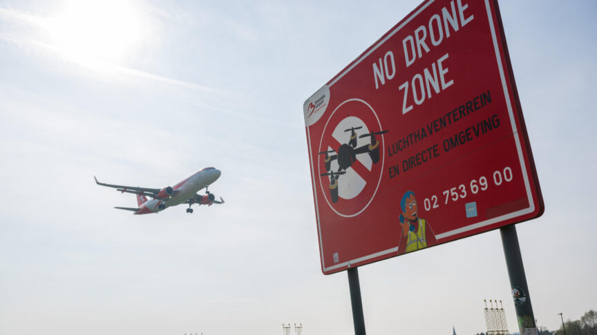 Ein Foto zeigt ein rotes 'No Drone Zone'-Schild im Vordergrund, ein Flugzeug landet im Hintergrund am Flughafen.