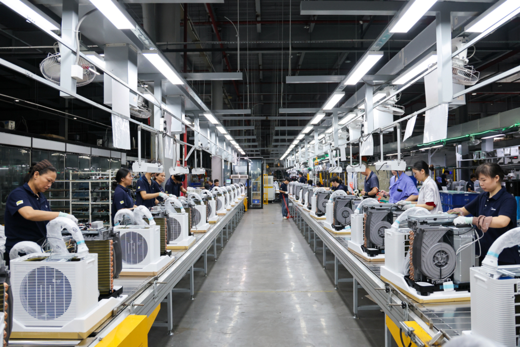 Ein Foto zeigt Arbeiter an einer Produktionslinie, die portable Split-Klimageräte in einer großen Fabrikhalle montieren.