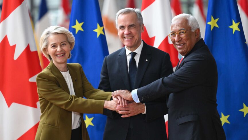 Ein Foto zeigt drei Personen, die sich die Hände übereinander legen vor kanadischen und EU-Flaggen in einem Saal.