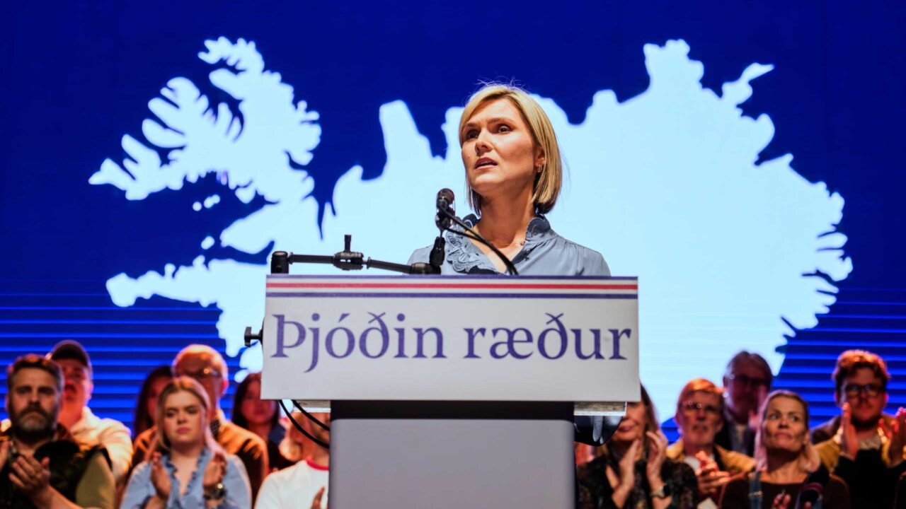 Ein Foto zeigt eine Frau am Rednerpult, sie spricht vor einem Publikum mit einer großen Karte der Insel im Hintergrund