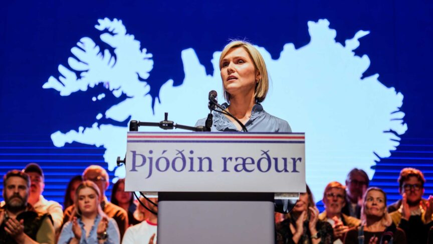 Ein Foto zeigt eine Frau am Rednerpult, sie spricht vor einem Publikum mit einer großen Karte der Insel im Hintergrund.