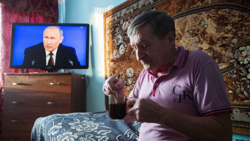 Ein Foto zeigt einen älteren Mann, der aus einem Glas Tee rührt, in einem Zimmer mit Fernseher und gemustertem Wandteppich.