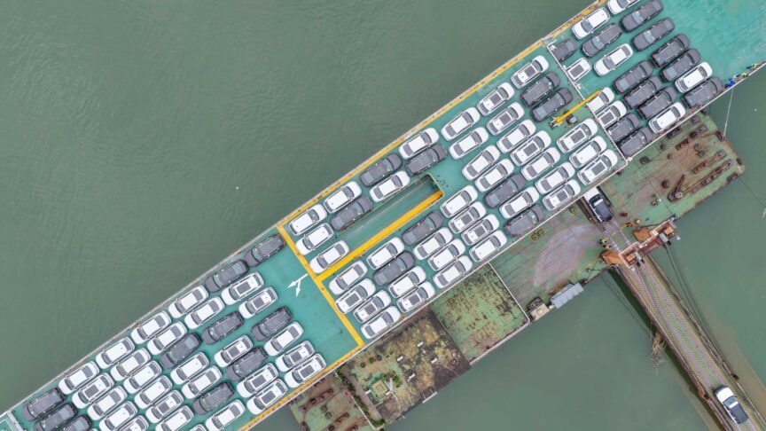 Ein Foto zeigt zahlreiche neue Autos beim Verladen auf ein Frachtschiff am Kai aus Vogelperspektive.