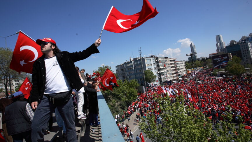 A man on a rooftop waves a Turkish flag above a square full of protestors