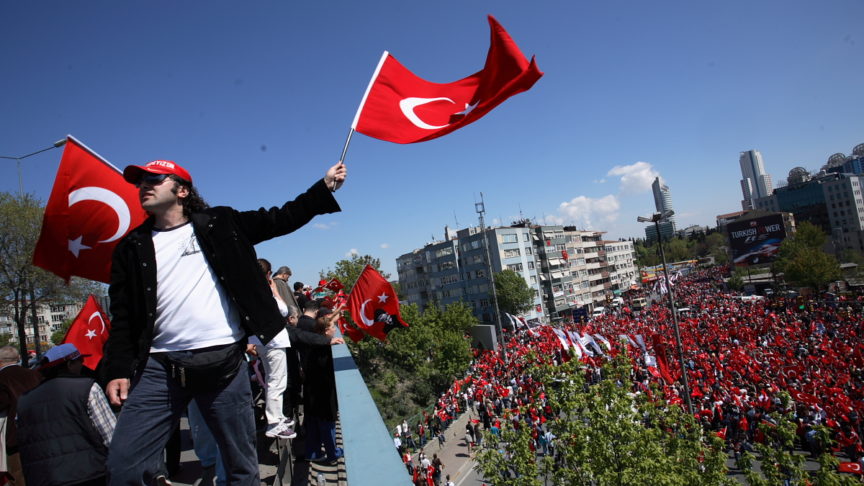 A man on a rooftop waves a Turkish flag above a square full of protestors